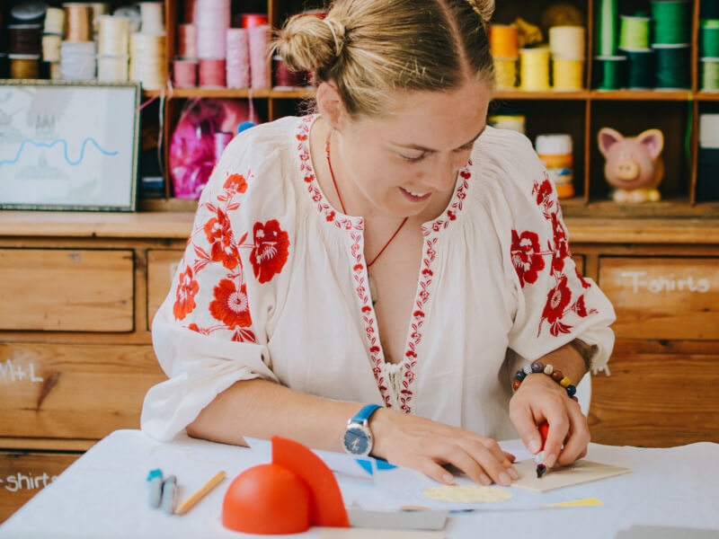 A woman is sitting at a table, carving a design into a block of lino